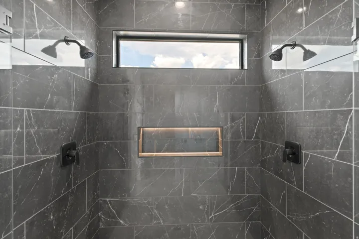 Dual matte black shower heads, dark marble-look tile, built-in shower niche with accent lighting, and transom window