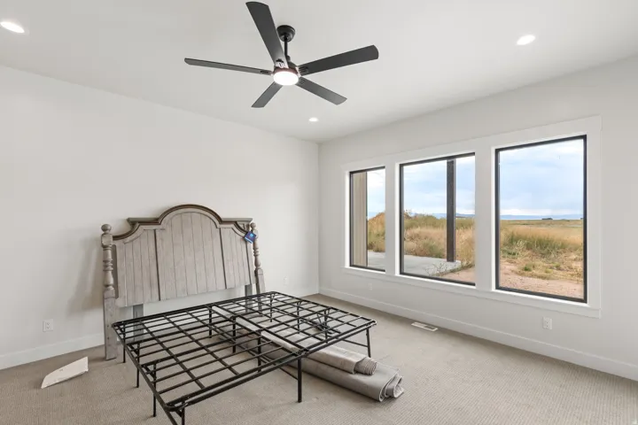 Recessed ceiling lighting, ceiling fan with integrated light, three large windows with black framing, light neutral-toned carpet flooring, and white painted walls