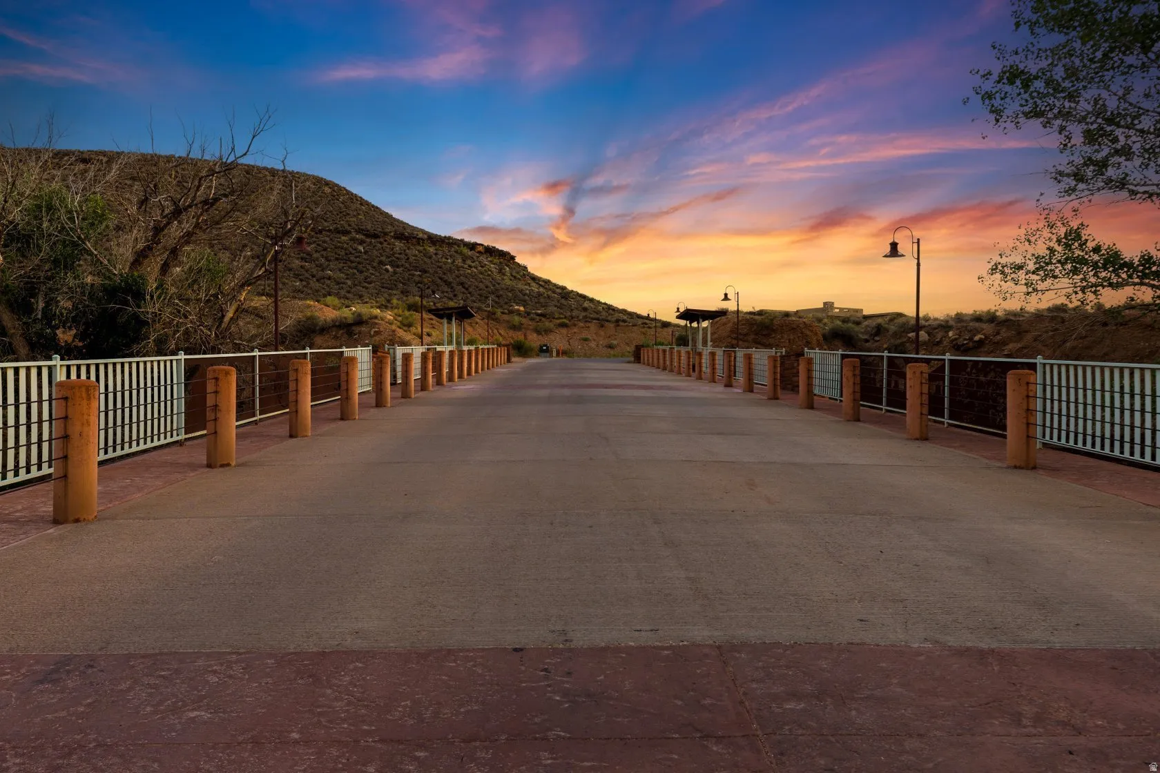 Paved pathway featuring white railings with wood-tone posts, street lamps, and a mountain backdrop