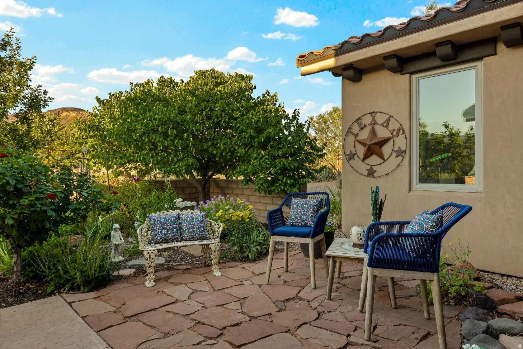 Flagstone patio featuring a stucco exterior with a window, decorative eaves, and a surrounding garden with mature trees and shrubs