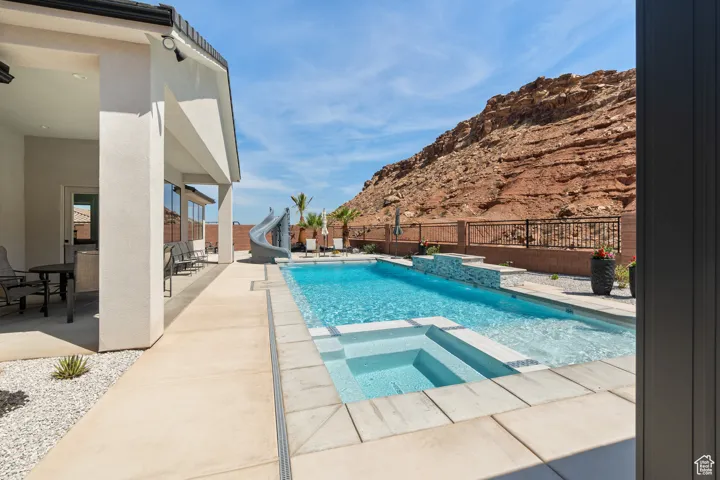 View of pool with a water slide, a patio, a fenced backyard, a pool with connected hot tub, and a mountain view