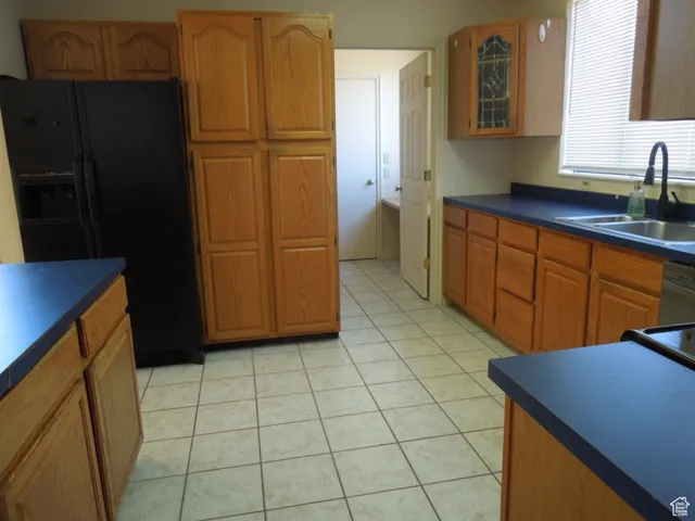 Kitchen featuring black fridge with ice dispenser, dark countertops, brown cabinetry, light tile patterned floors, and dishwasher