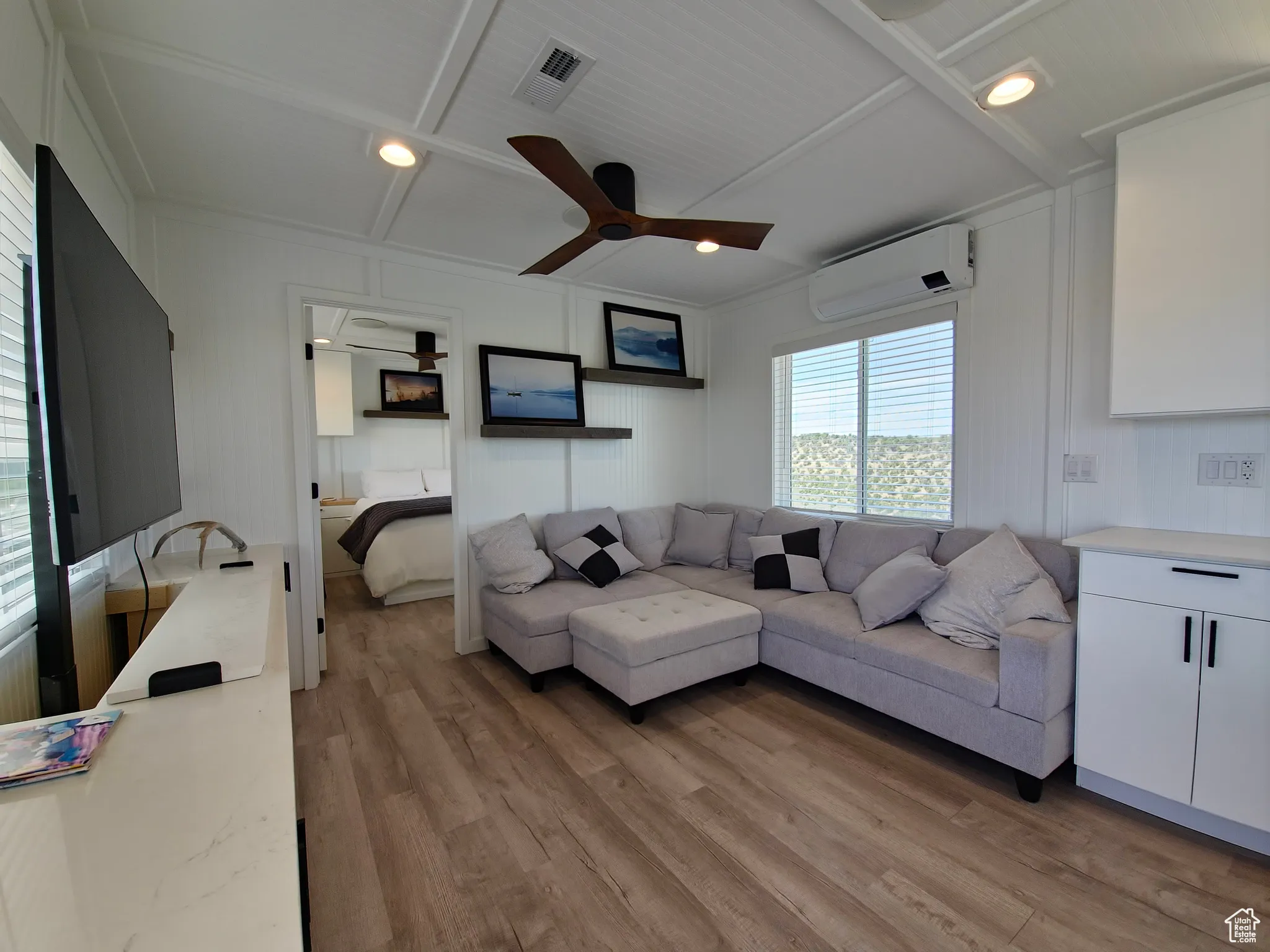 Living area with a wall mounted air conditioner, a ceiling fan, light wood finished floors, a decorative wall, and beamed ceiling