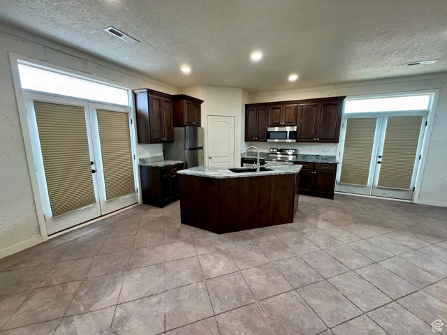 Kitchen featuring appliances with stainless steel finishes, a textured ceiling, dark brown cabinets, french doors, and a kitchen island with sink