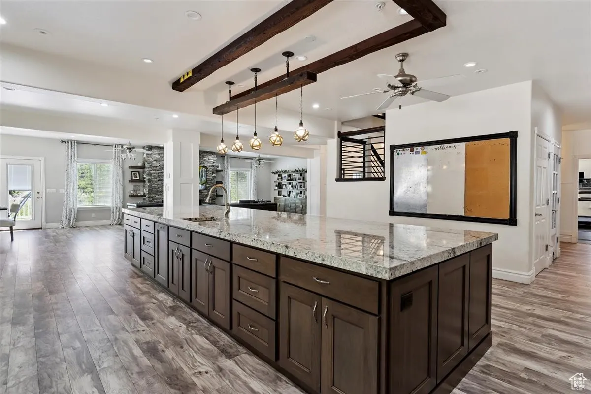 Kitchen featuring light wood finished floors, dark brown cabinetry, a ceiling fan, beamed ceiling, and light stone countertops