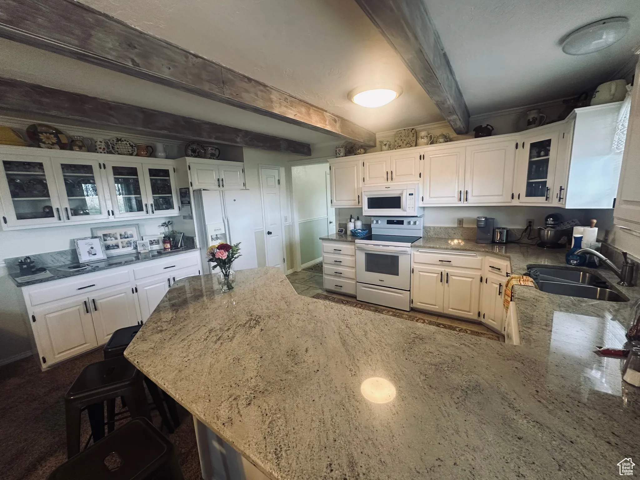 Kitchen with white appliances, light stone counters, white cabinetry, beamed ceiling, and a peninsula