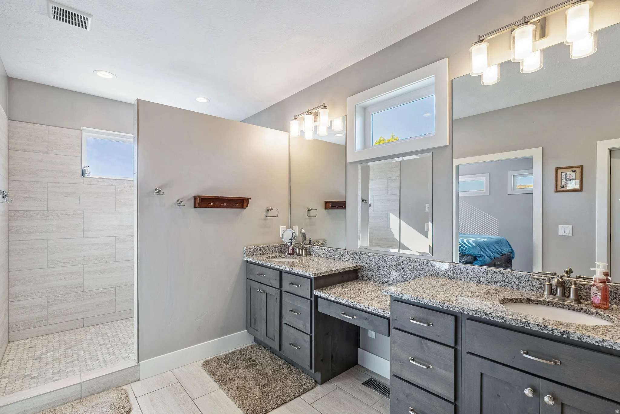 Spacious bathroom featuring a walk-in shower with light-toned tile, dual vanities with stone countertops, and dark wood cabinetry