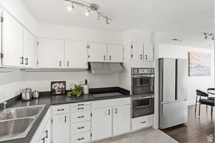 Kitchen with stainless steel fridge, track lighting, multiple ovens, dark countertops, and white cabinetry