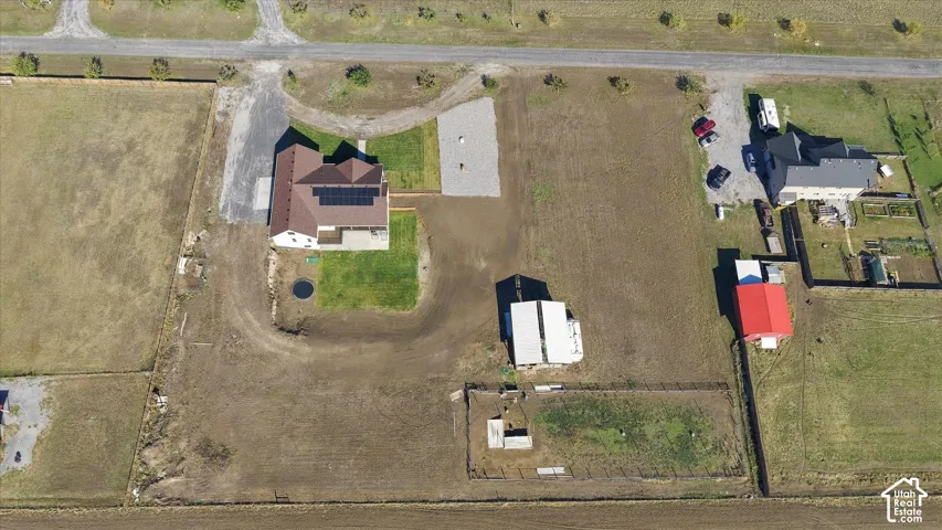 Aerial view of a multi-acre property featuring a residential structure with rooftop solar panels, a detached metal-sided outbuilding, a gravel parking area, and fenced livestock enclosures