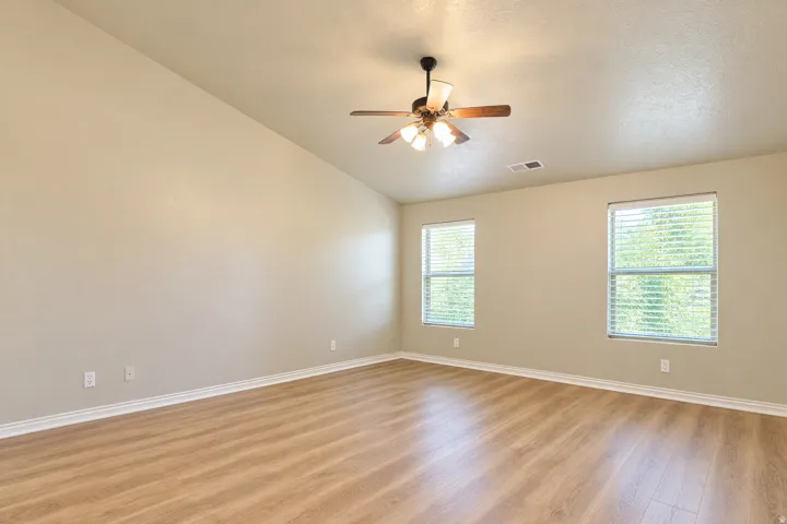 Vaulted ceiling, wood-finish flooring, two windows with blinds, ceiling fan with light fixture, neutral wall color