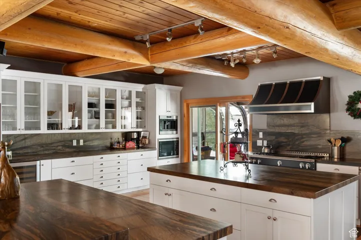 Kitchen featuring butcher block countertops, extractor fan, backsplash, glass insert cabinets, and a wooden ceiling with exposed beams