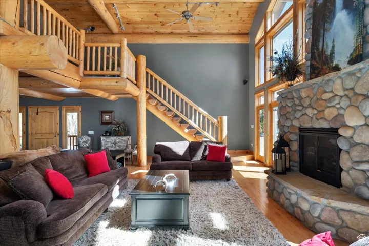 Living room with wood finished floors, stairway, a stone fireplace, a wood ceiling with exposed beams, and a ceiling fan