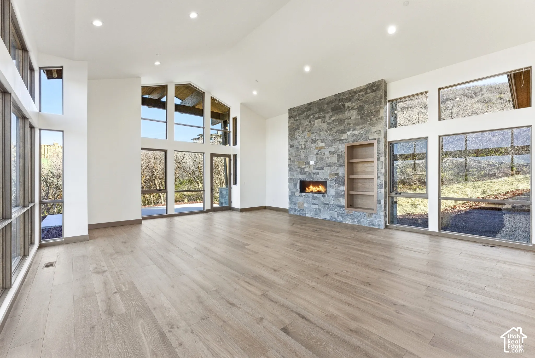 Unfurnished living room with a high ceiling, a stone fireplace, healthy amount of natural light, light wood finished floors, and recessed lighting