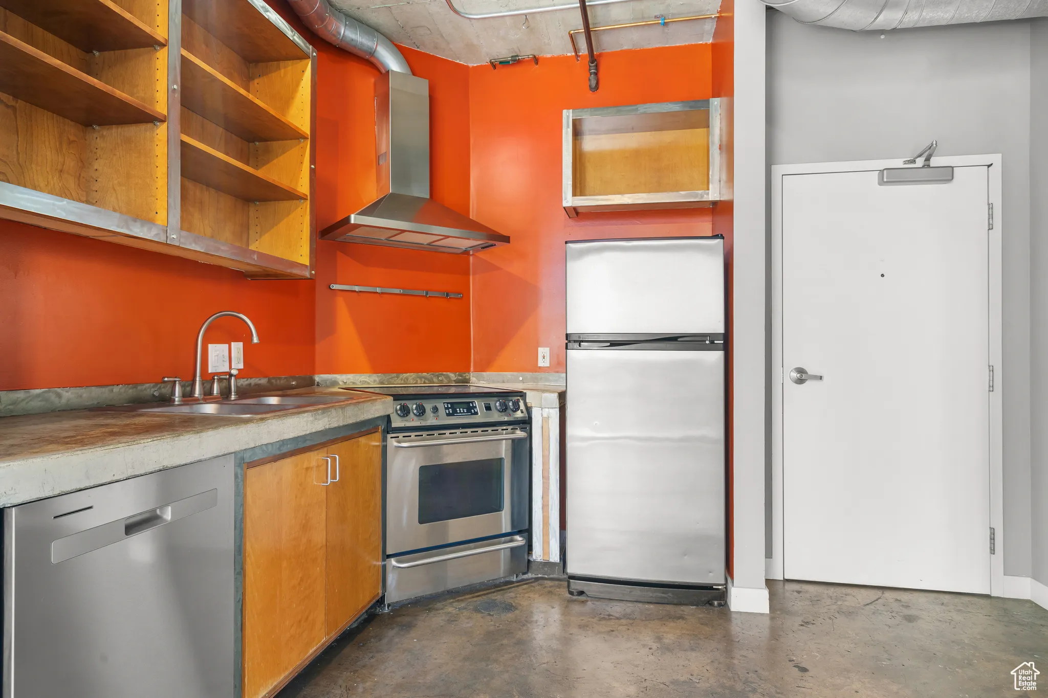 Kitchen with wall chimney range hood, appliances with stainless steel finishes, concrete flooring, open shelves, and brown cabinetry