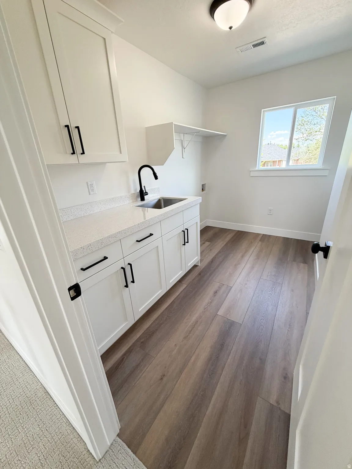 Utility room featuring wood-finish flooring, white cabinetry with black hardware, a stainless steel sink with a matte black faucet, a quartz-look countertop, and a window