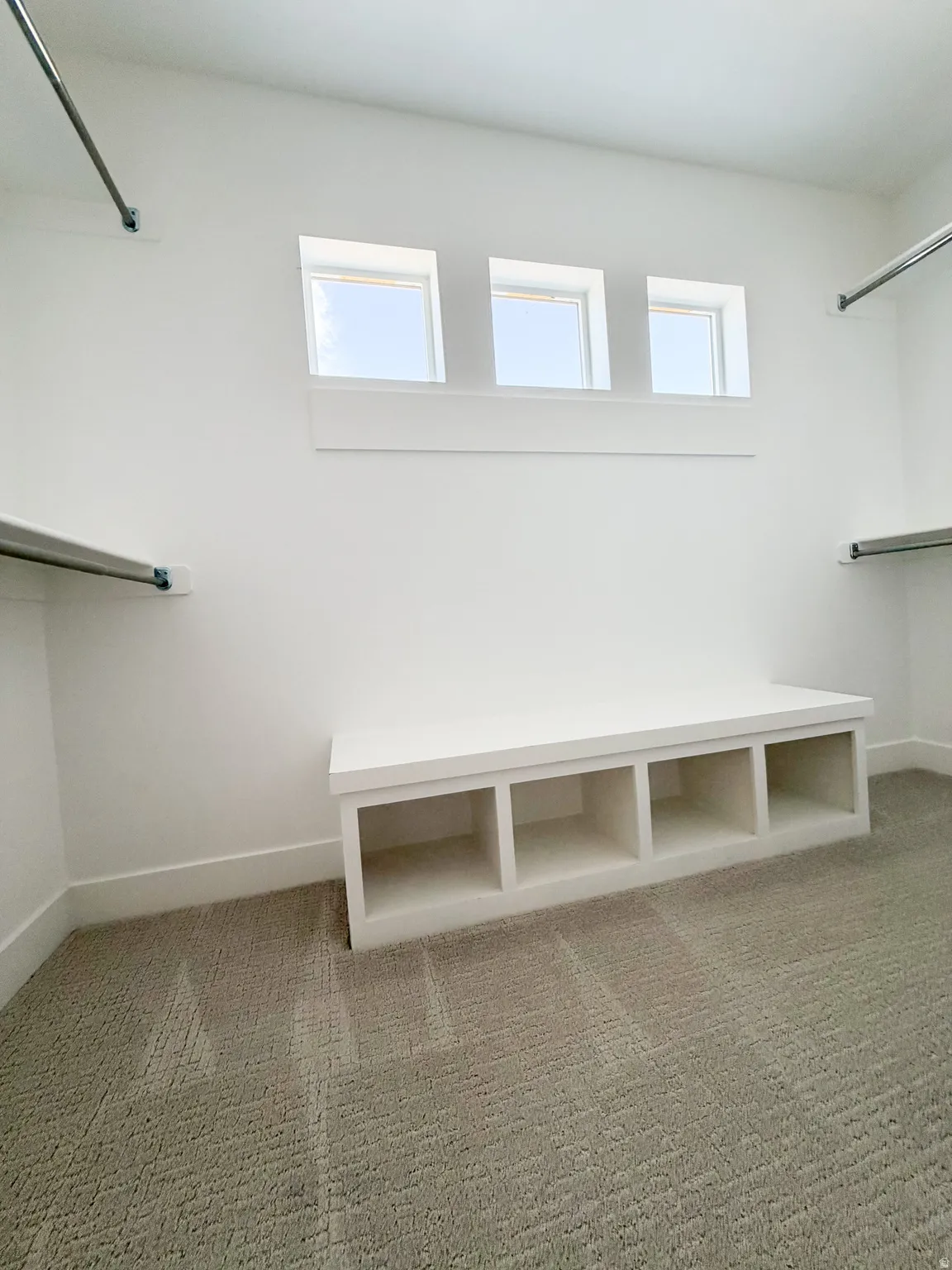 Spacious walk-in closet featuring light-toned carpeting, ample built-in shelving, three high windows providing natural illumination, and a built-in storage bench with four compartments