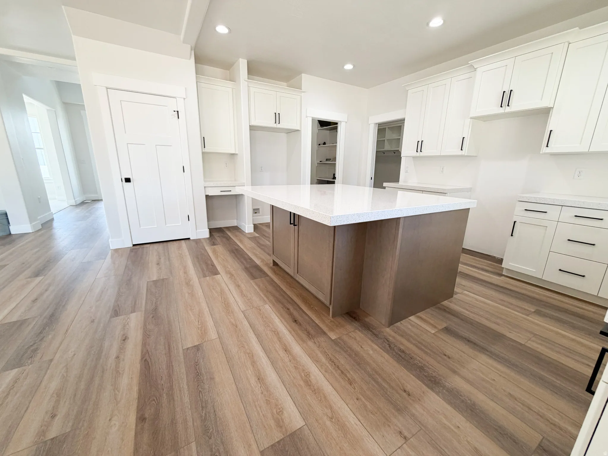 Modern kitchen featuring a large island with a light-colored countertop, wood-finish flooring, white upper and lower cabinetry, and black hardware