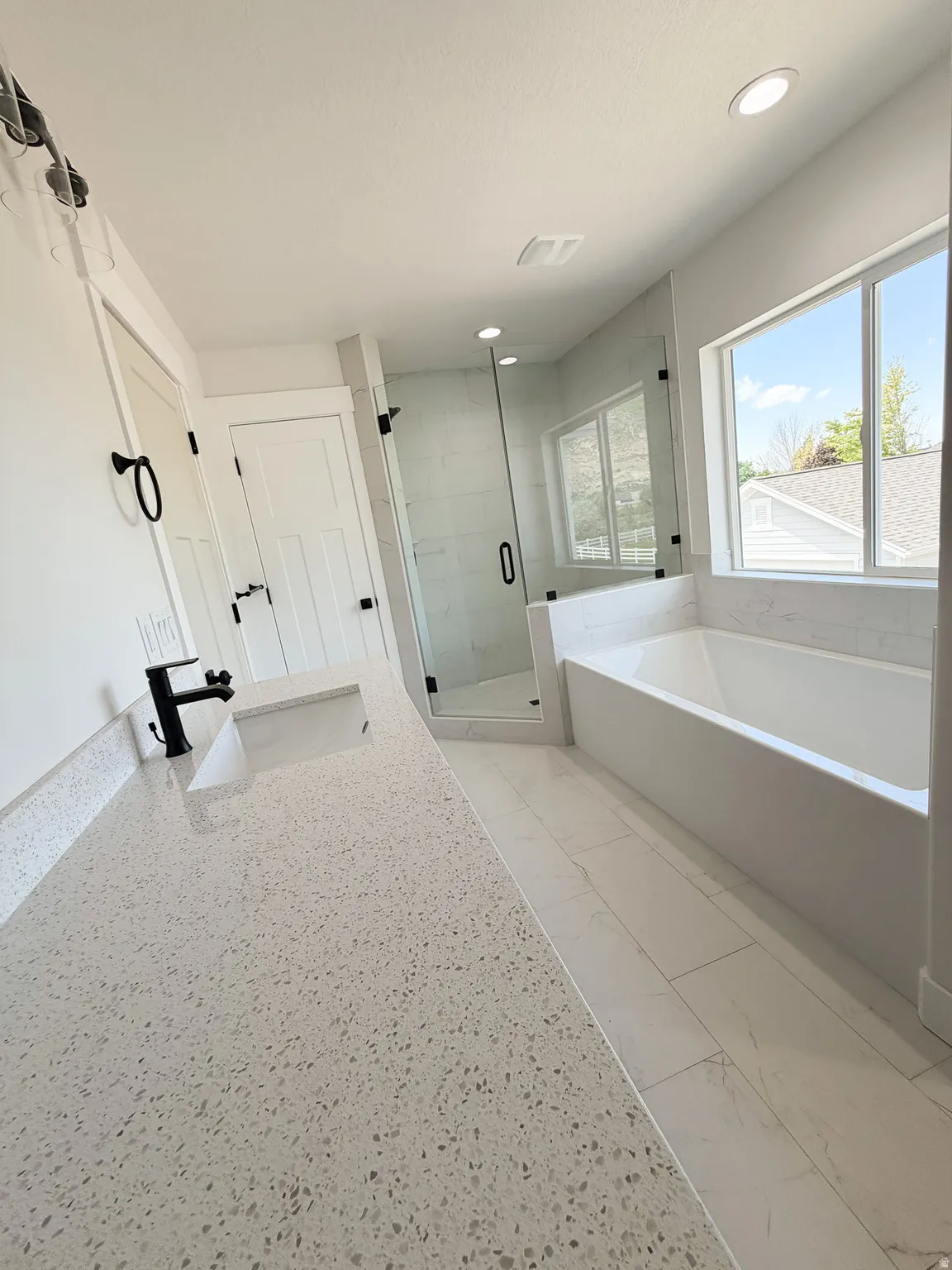 Contemporary bathroom featuring a spacious white countertop with an integrated sink and black faucet, a large soaking tub with a tiled surround, a glass-enclosed shower with white subway tile, a wide window, and recessed ceiling lighting