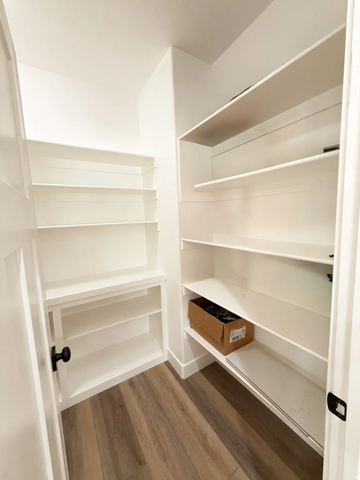 Walk-in pantry featuring extensive white shelving, wood-finish flooring, and a modern black door handle