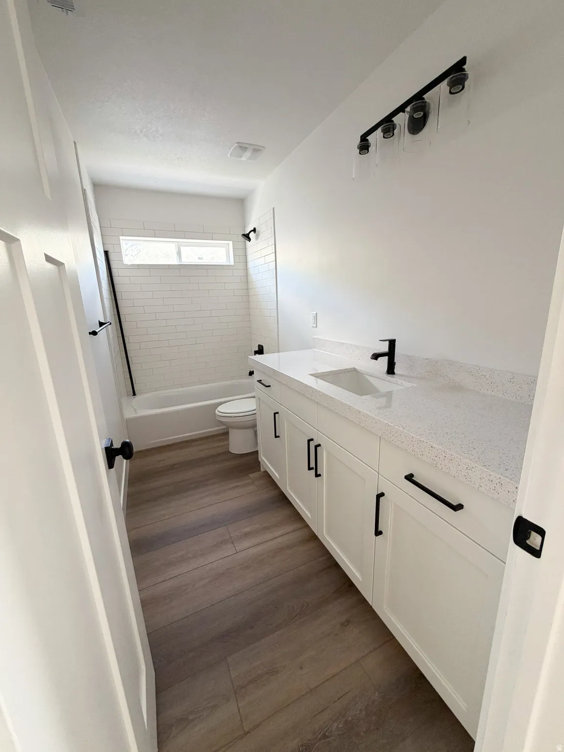 Bathroom featuring a white subway tile tub surround, white vanity with black hardware, white countertop, wood-finish flooring, and a black-finish vanity light fixture