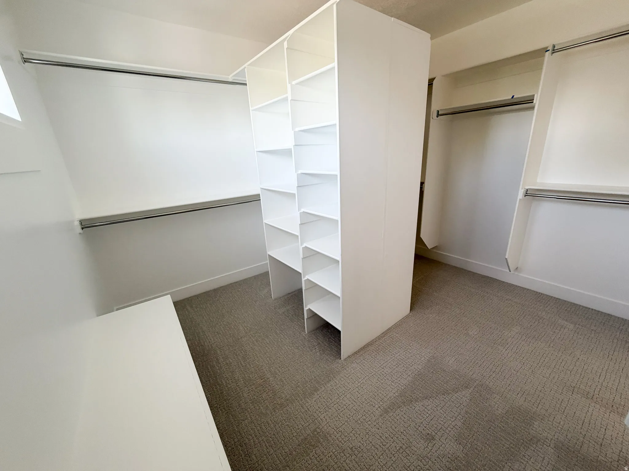 Spacious closet featuring a central shelving unit, multiple hanging rods, white walls, light-colored carpet, and natural light