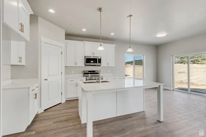 Kitchen featuring wood finished floors, appliances with stainless steel finishes, a kitchen island with sink, light countertops, and white cabinetry