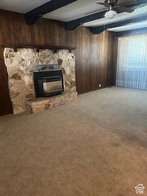 Unfurnished living room featuring beamed ceiling, wooden walls, light colored carpet, a ceiling fan, and a stone fireplace