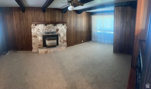Unfurnished living room with wood walls, beam ceiling, a ceiling fan, light carpet, and a stone fireplace