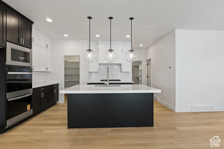 Kitchen with tasteful backsplash, light countertops, a kitchen island with sink, double oven, and light wood-style floors