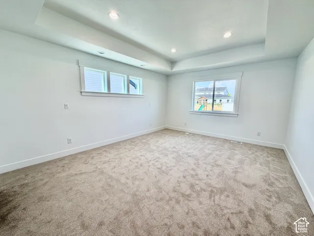Spacious interior room featuring a tray ceiling with recessed lighting, plush carpeting, multiple windows for ambient light, and white trim