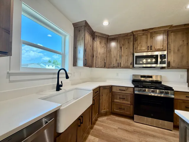 Kitchen featuring wood-finish cabinetry, white countertops, stainless steel appliances, a farmhouse sink with a matte black faucet, and wood-finish flooring