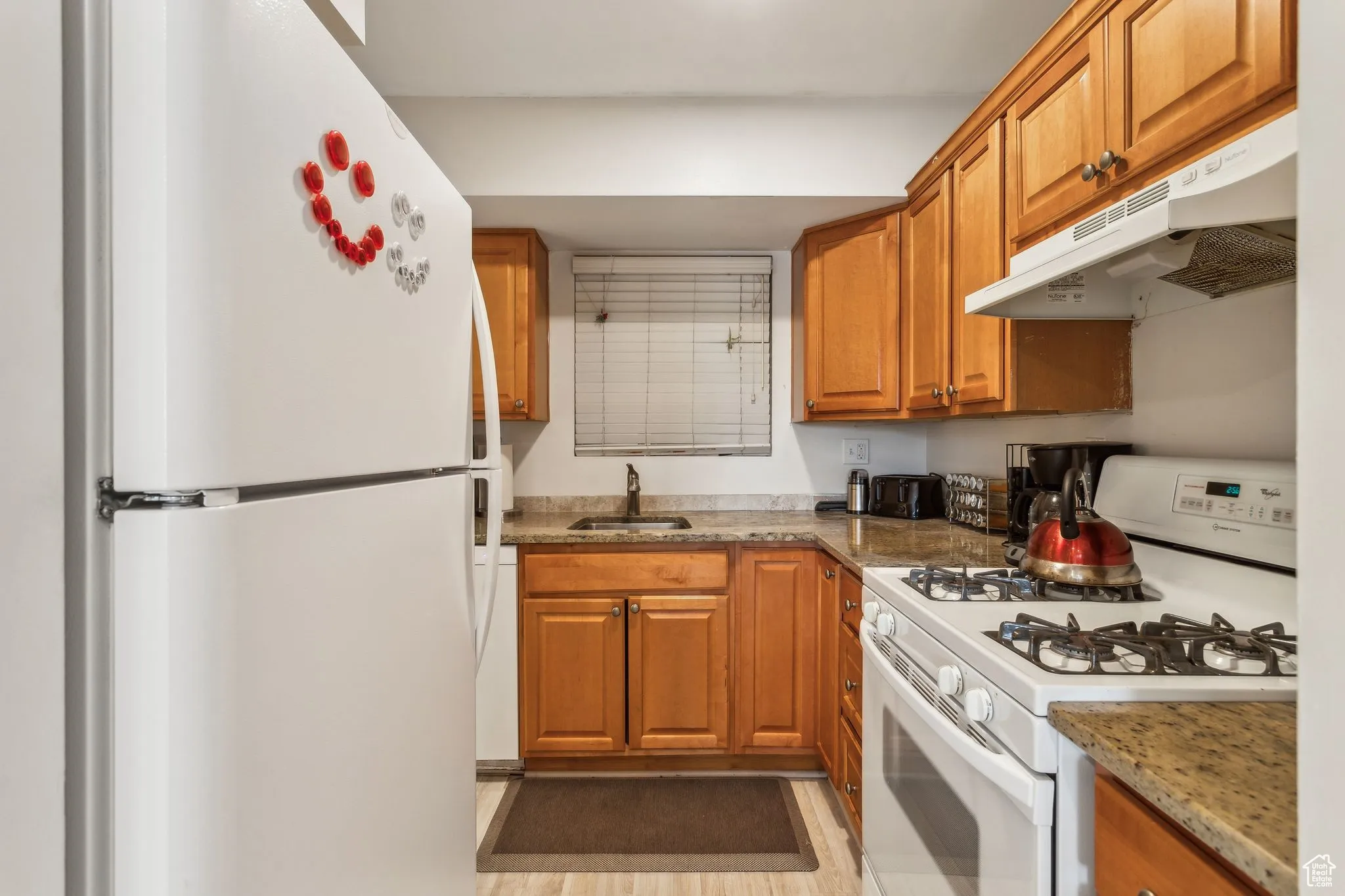 Kitchen featuring white appliances, under cabinet range hood, brown cabinets, light stone countertops, and light wood-type flooring