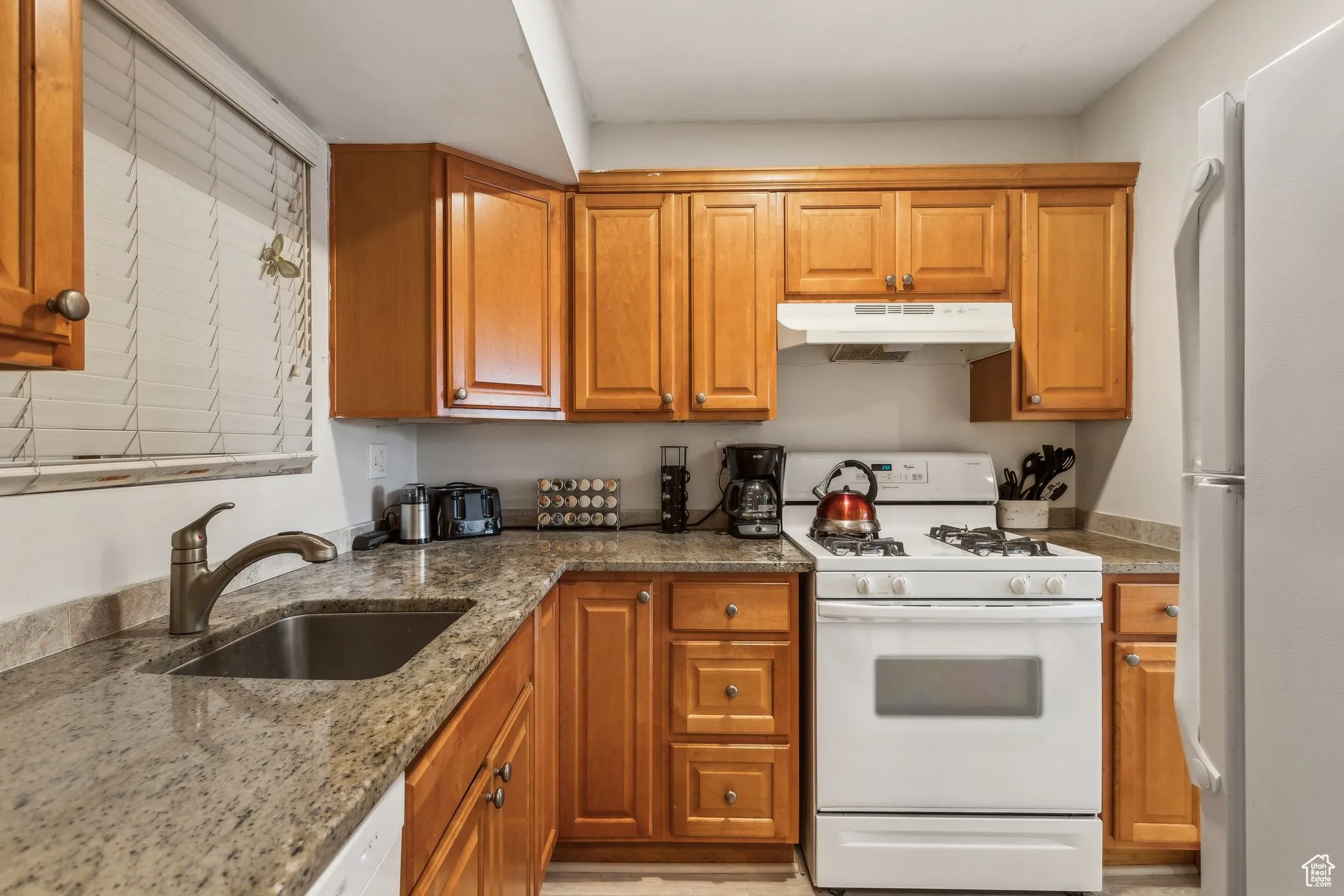 Kitchen with white appliances, under cabinet range hood, stone counters, and brown cabinets