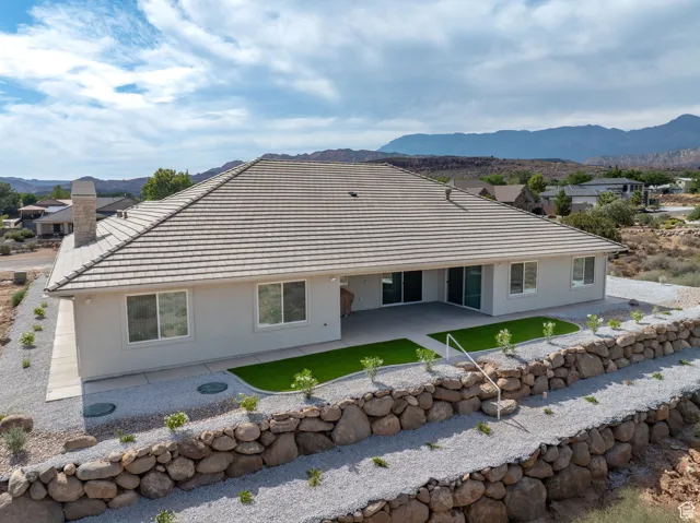 Rear view of property featuring a patio area, stucco siding, terraced rear with stone retaining walls and step down ladders, and a chimney to the wood-burning fireplace in the family room.  Concrete sidewalks surround the home.