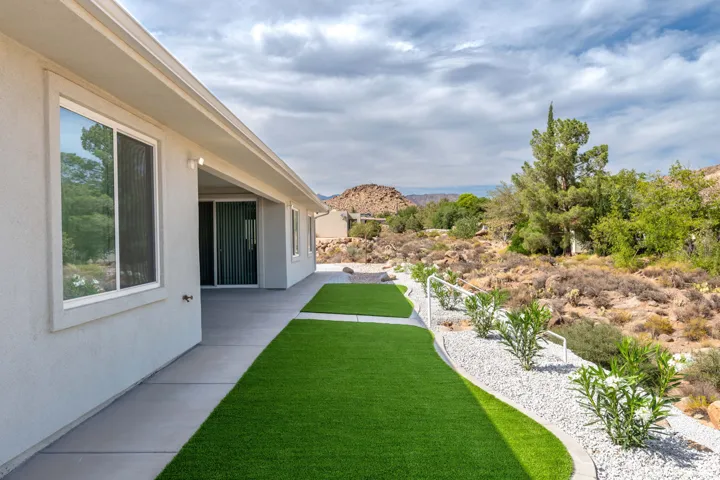 View of grassy artificial turf yard featuring a patio area & natural terrain.  Concrete sidewalks surround the home.