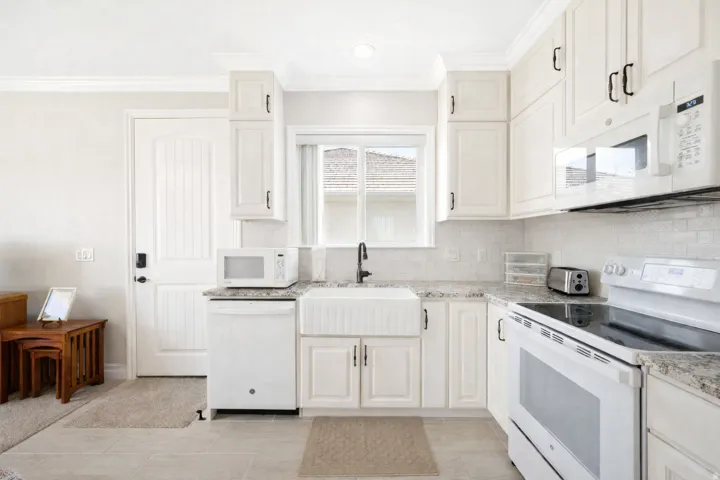 Apartment kitchen featuring white appliances, light stone counters, backsplash, crown molding, and white cabinetry