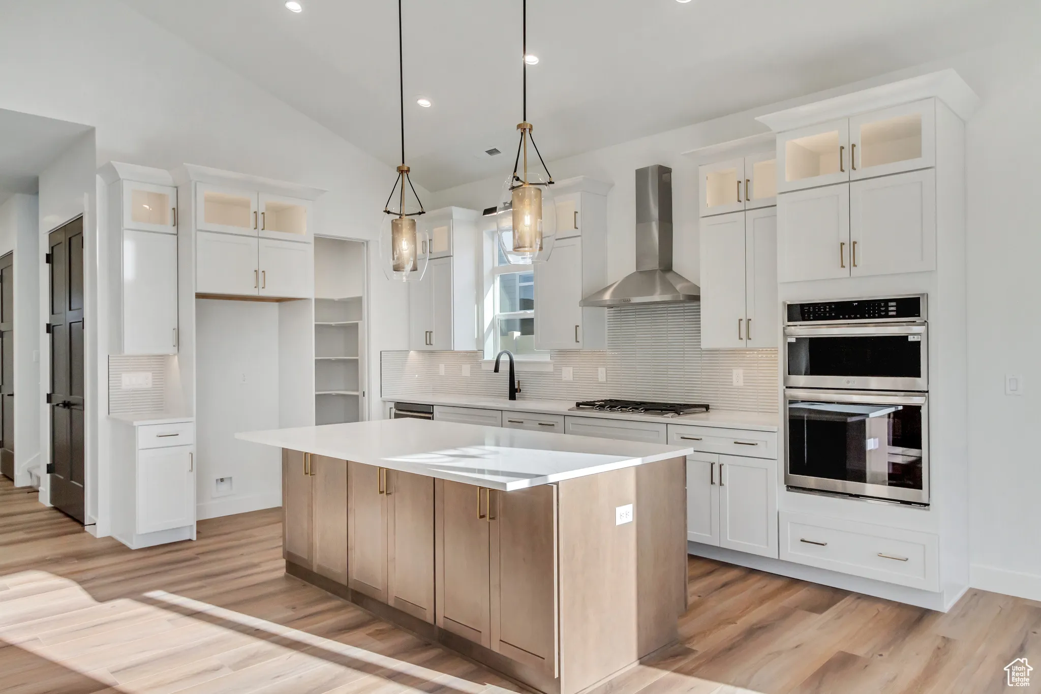 Kitchen with glass insert cabinets, backsplash, decorative light fixtures, wall chimney range hood, and lofted ceiling