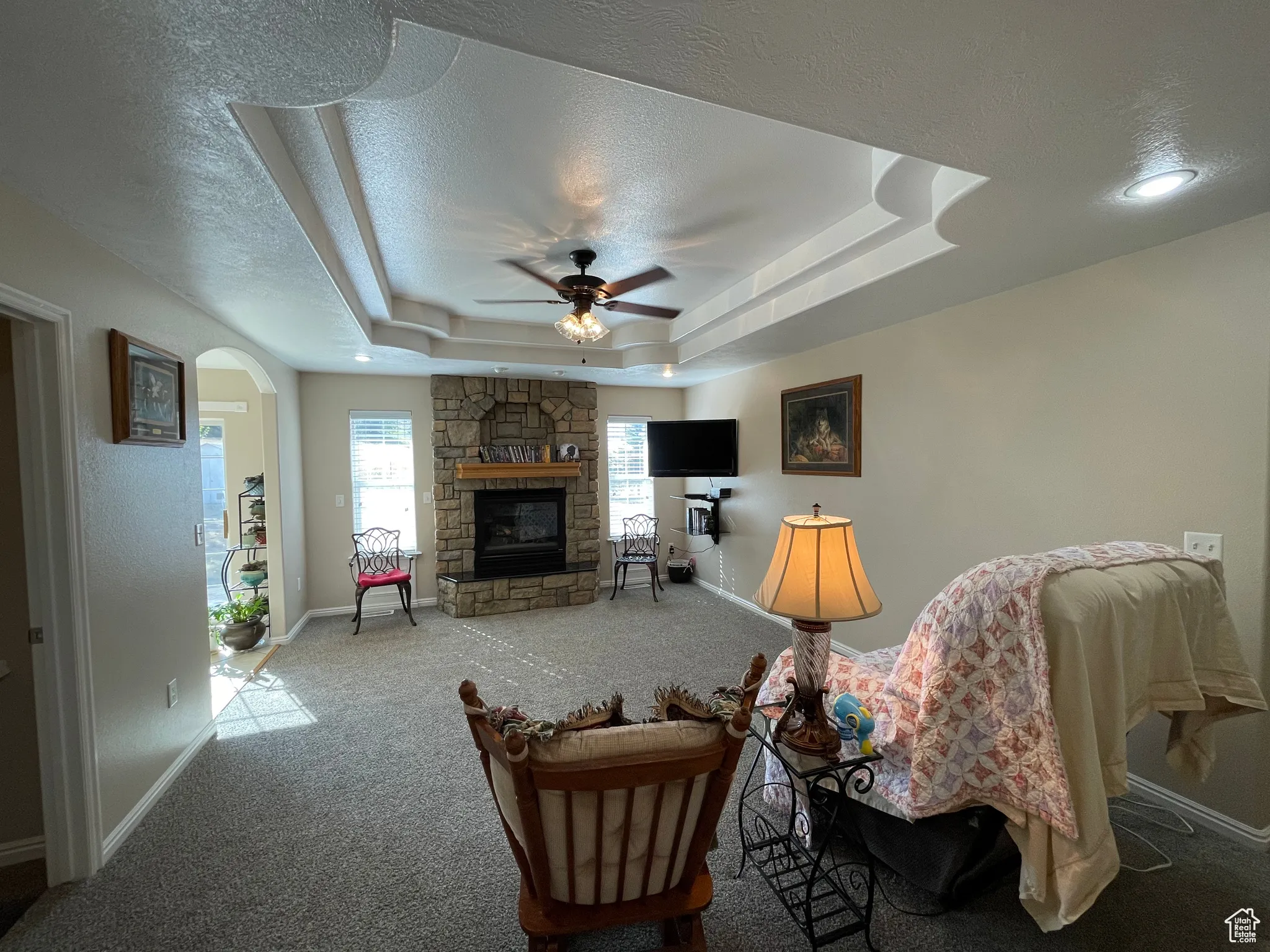 Carpeted living area with arched walkways, a stone fireplace, a raised ceiling, a textured ceiling, and ceiling fan