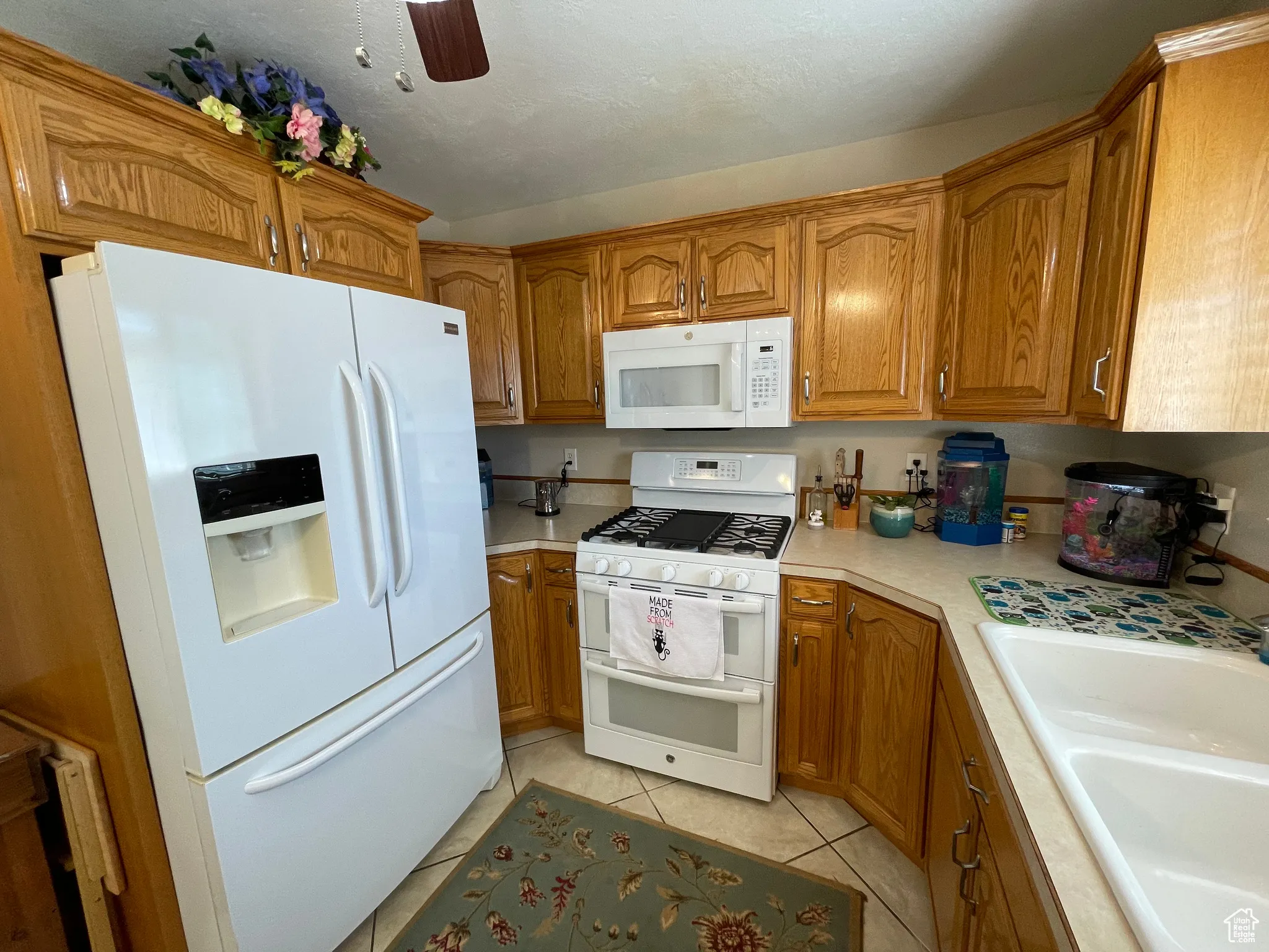 Kitchen with white appliances, brown cabinets, light countertops, light tile patterned flooring, and ceiling fan