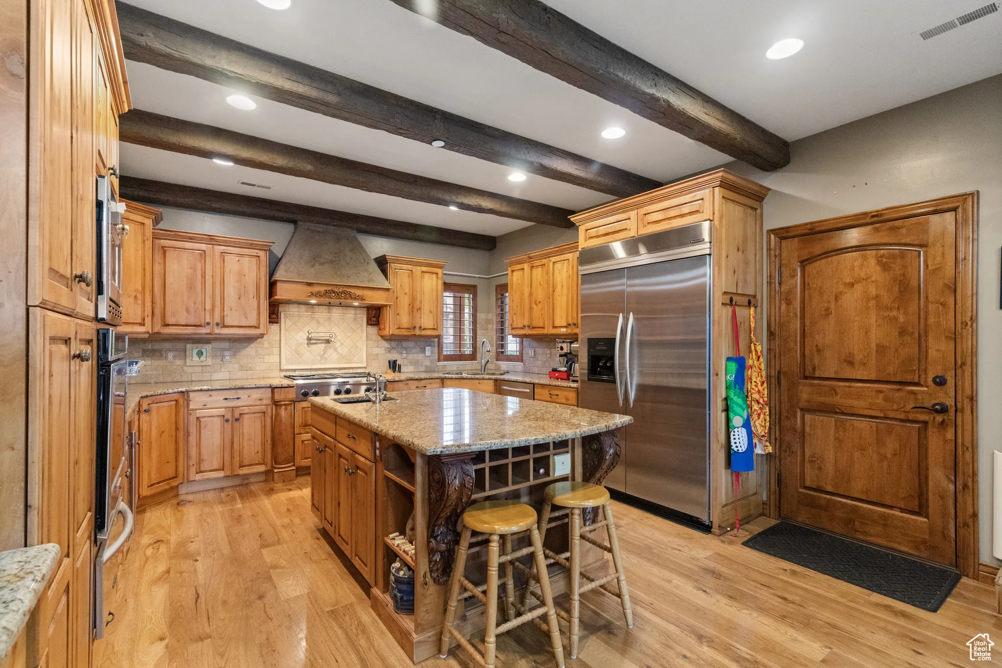 Kitchen with tasteful backsplash, light wood-style flooring, appliances with stainless steel finishes, beamed ceiling, and an island with sink