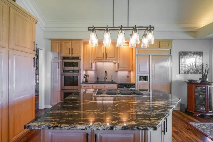 Kitchen with dark stone countertops, dark wood-type flooring, decorative backsplash, decorative light fixtures, and ornamental molding