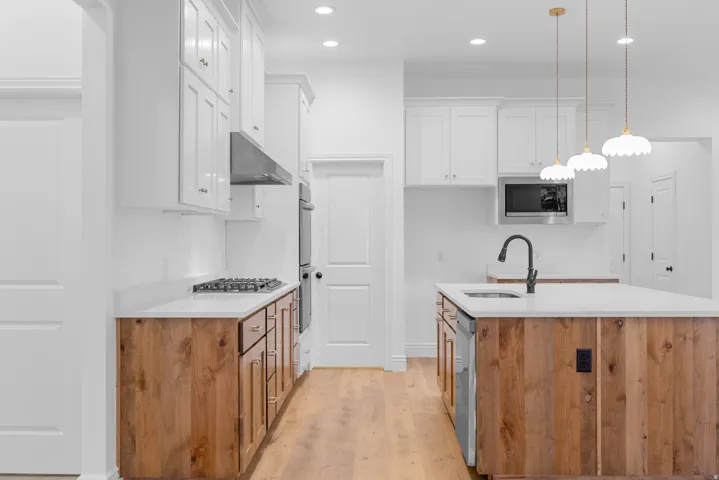 Kitchen featuring an island with sink, light wood finished floors, stainless steel appliances, light stone countertops, and decorative light fixtures
