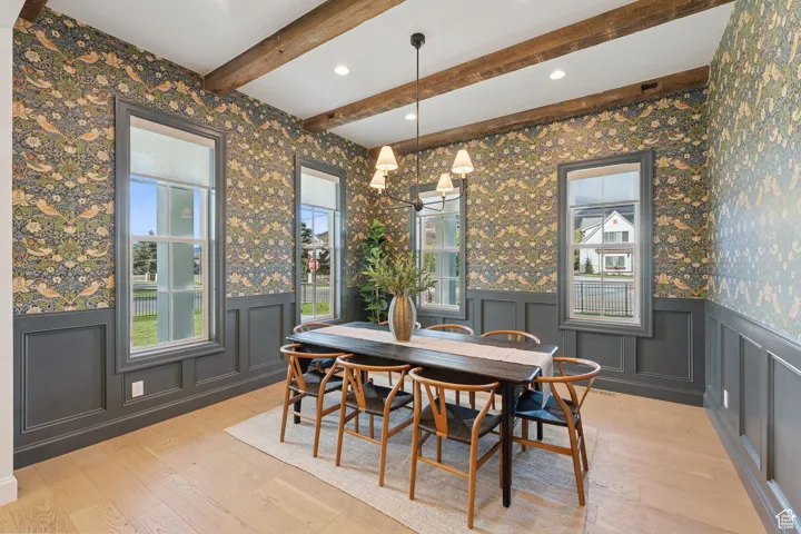 Dining space featuring wallpapered walls, wainscoting, a chandelier, beamed ceiling, and light wood-style floors