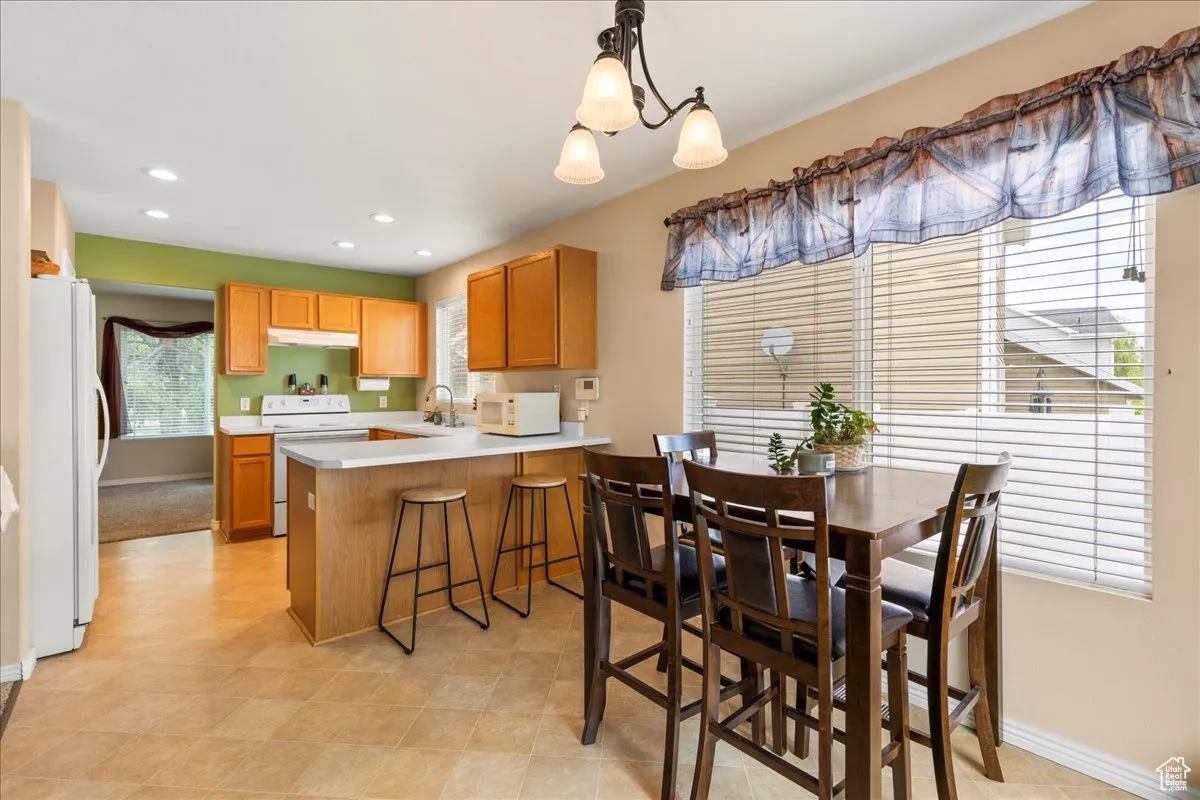 Kitchen with light countertops, a peninsula, white appliances, hanging light fixtures, and a kitchen bar