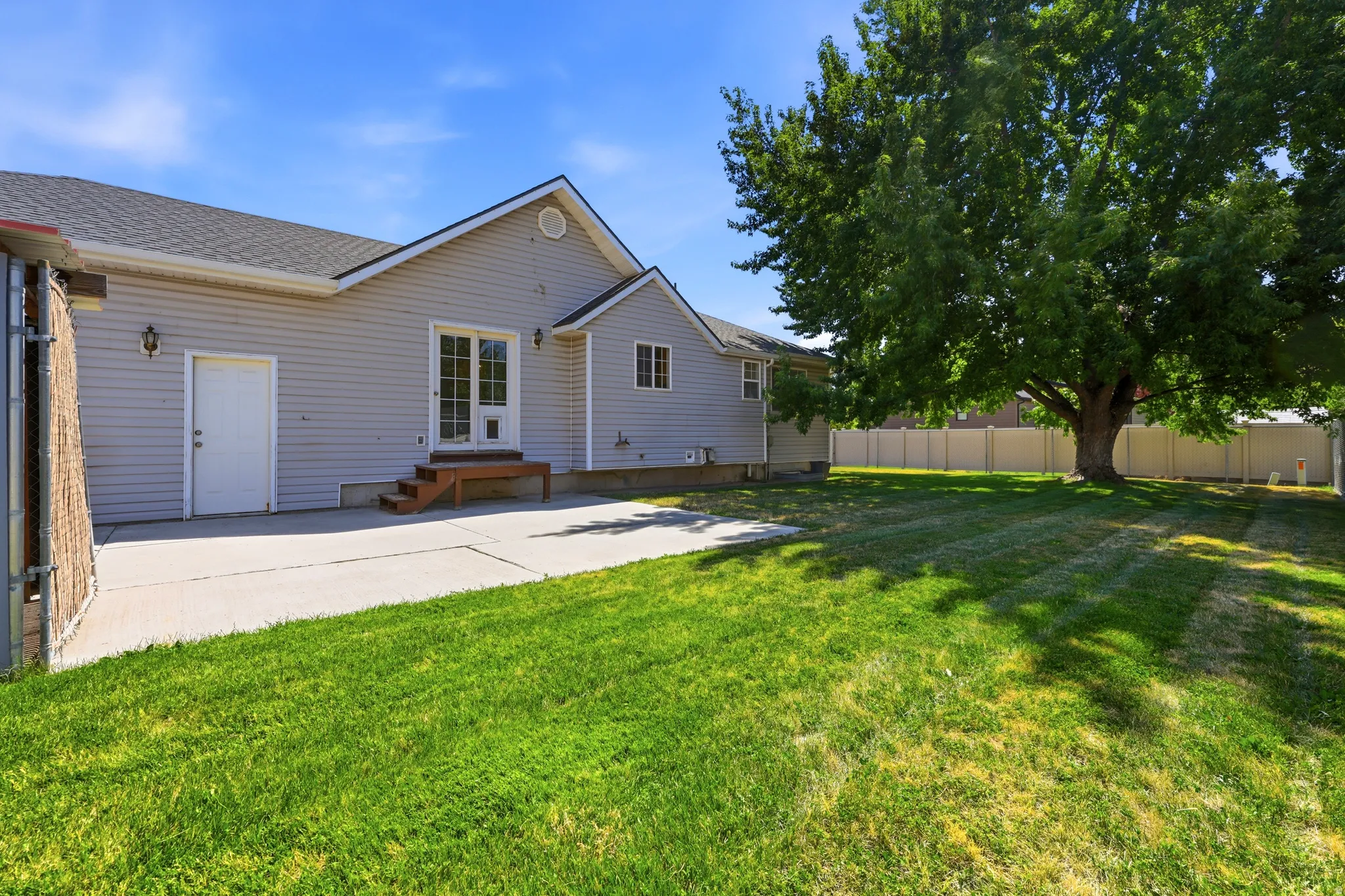 Expansive green lawn with a mature shade tree, concrete patio, and a light-colored vinyl fence