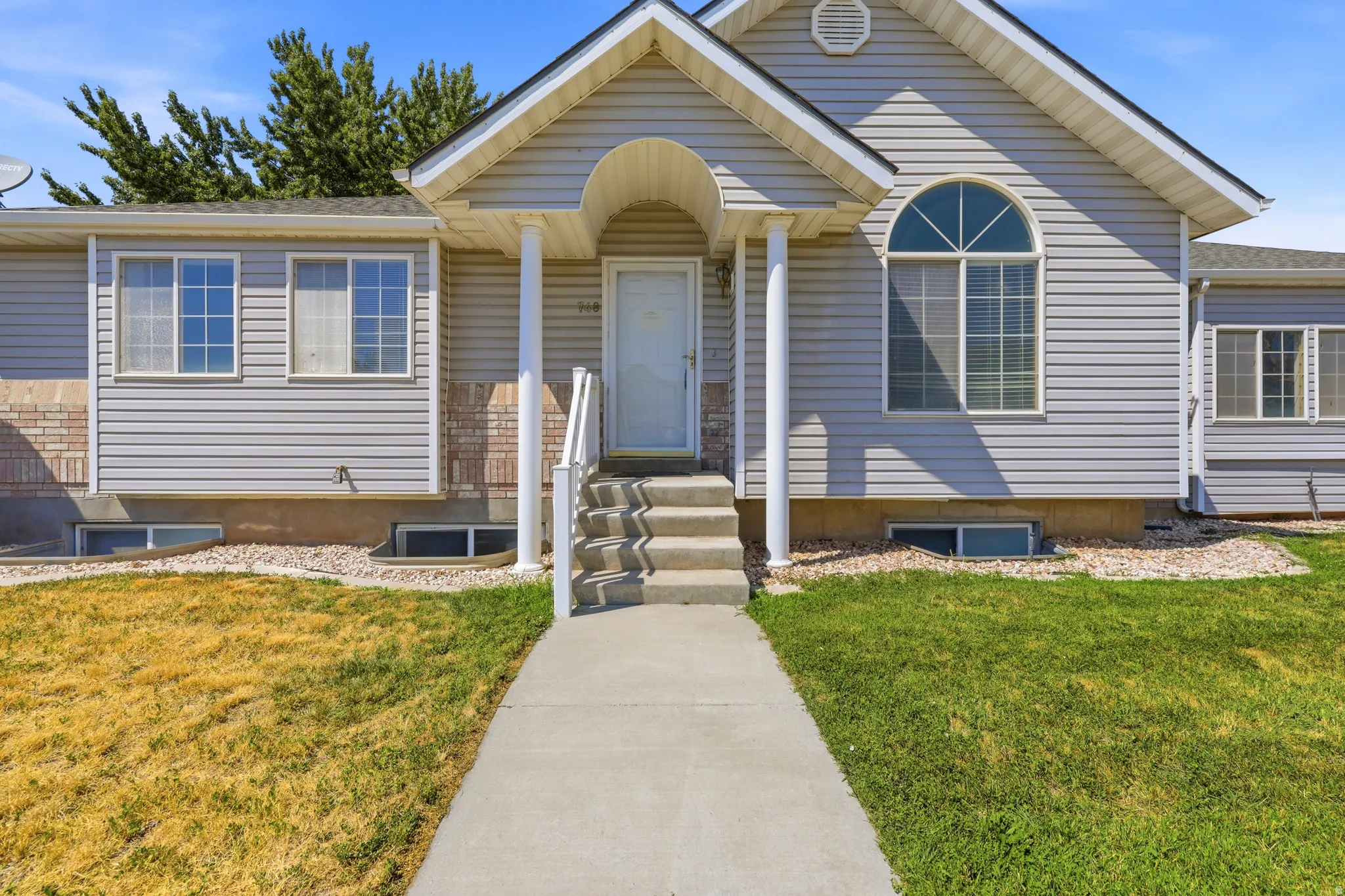 Light gray siding exterior with a covered entry, an arched window, a concrete walkway, and a partial brick facade