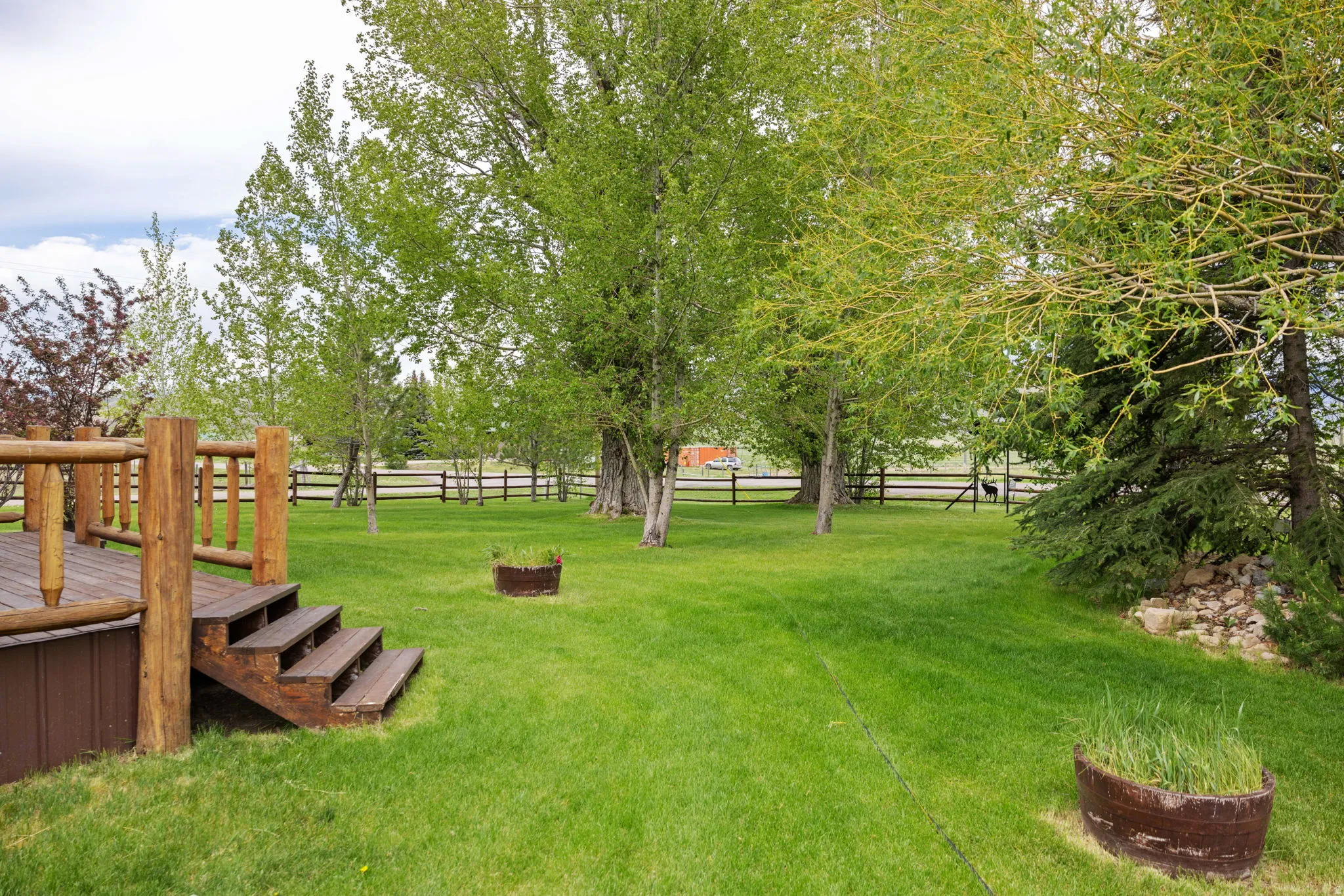 Spacious green lawn featuring a wooden deck with log railings and steps, bordered by mature trees and a distant wooden fence