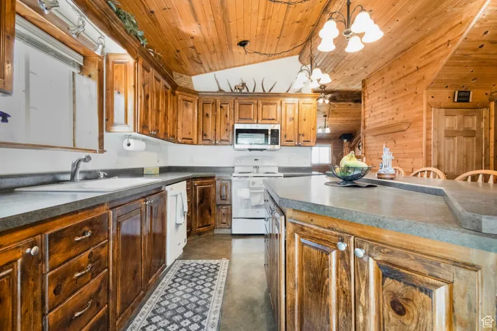 Kitchen featuring finished concrete flooring, white appliances, vaulted ceiling, brown cabinetry, and decorative light fixtures
