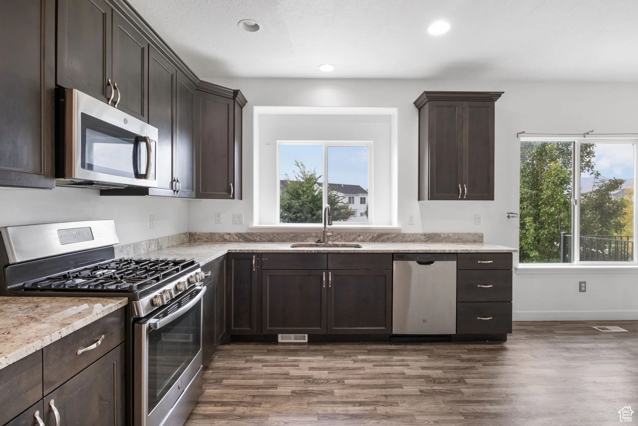 Kitchen with dark brown cabinetry, stainless steel appliances, dark wood-style flooring, light stone counters, and recessed lighting