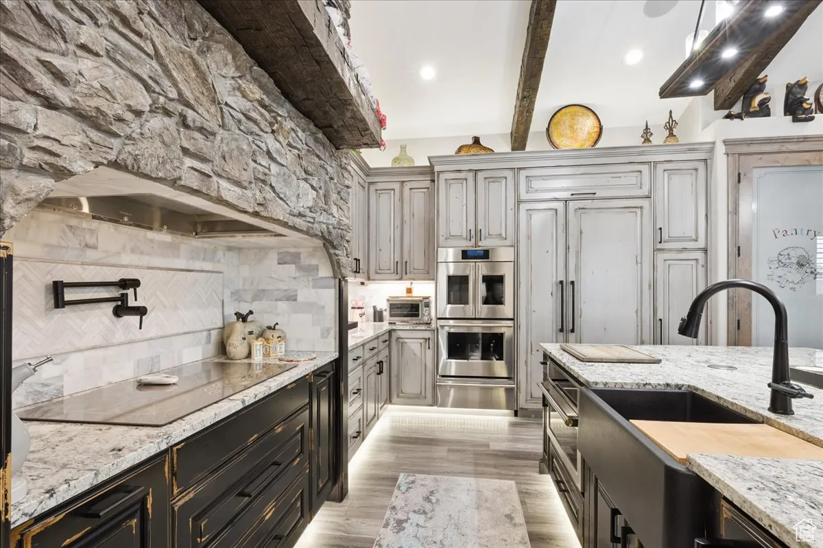 Kitchen featuring light stone countertops, double oven, decorative backsplash, beamed ceiling, and light wood-style flooring